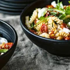 Zwei Schalen mit Nudelsalat mit getrockneten Tomaten, Pinienkernen und Rucola. Ein Löffel liegt in der linken Schale.