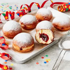Berliner Rezept Original: Sieben luftige Krapfen mit Marmeladenfüllung, bestäubt mit Puderzucker, auf einem Blech, umgeben von bunten Party-Girlanden und Konfetti.