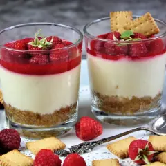 Zwei Gläser mit Himbeer-Dessert: eine Schicht Keksbrösel, cremige Masse und frische Himbeeren mit Himbeersauce.