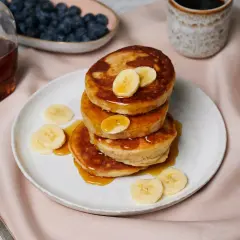 Ein Stapel goldbrauner Bananen-Pancakes, beträufelt mit Sirup und garniert mit Bananenscheiben. Daneben Blaubeeren, Ahornsirup und Kaffee.