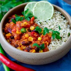 Herzhaftes Chili sin Carne mit Reis, garniert mit frischer Petersilie und Limettenspalten. Rote und grüne Chilis liegen daneben.