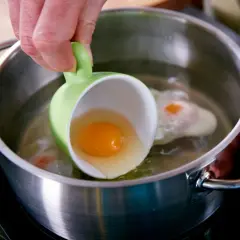 Ein rohes Ei wird aus einer grünen Tasse in kochendes Wasser gegossen. Ein Schritt für ein perfektes verlorene Eier Rezept.