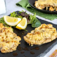 Zwei Portionen überbackenes Steak au four mit geschmolzenem Käse, serviert mit Salat und Zitronenspalten auf einem schwarzen Schiefer.