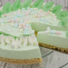 Eine helle, cremige Philadelphia Torte mit Waldmeistergeschmack, dekoriert mit Marshmallows und grünen Blättern. Ein Stück wird gerade serviert.