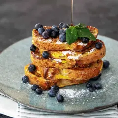 Ein Stapel goldbrauner French Toast, garniert mit Blaubeeren und Puderzucker. Ein dünner Strahl Sirup fließt darüber.