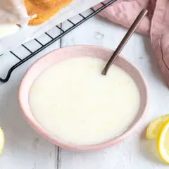 Zitronenglasur selber machen: cremige Glasur in einer Schale, daneben Zitronenspalten und ein Stück Kuchen auf einem Gitter.
