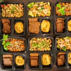 Vorbereitete Mahlzeiten in schwarzen Behältern: Pasta mit Brokkoli, Linsen mit Sonnenblumenkernen und Bohnen-Eintopf. Dazu Flachbrot und Hummus – perfekt für low carb high protein.