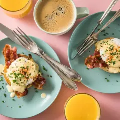 Zwei Teller mit Eggs Benedict, dazu Kaffee und Orangensaft auf rosa Untergrund.