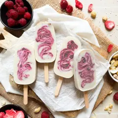Vier selbstgemachte Eis am Stiel mit Himbeermuster, umgeben von frischen Himbeeren, Cashews und gefriergetrockneten Erdbeeren.