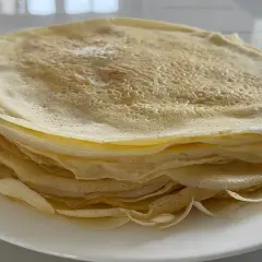 Ein Stapel goldbrauner Crêpes, frisch zubereitet aus dem Teig, liegt auf einem weißen Teller.
