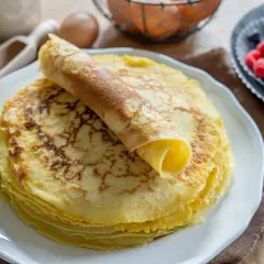 Ein Stapel goldbrauner Pfannkuchen, einer davon aufgerollt, wartet auf ein leckeres Pfannkuchen Rezept. Beeren und Eier im Hintergrund.