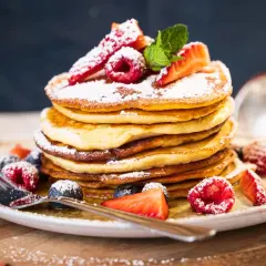Ein Stapel fluffiger amerikanischer Pancakes, bestäubt mit Puderzucker und garniert mit frischen Beeren und Minze. Ein köstliches american pancakes rezept für den perfekten Brunch.