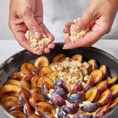 Hände streuen Streusel über saftige Pflaumen in einer Springform. Ein köstlicher saftiger Pflaumenkuchen entsteht.