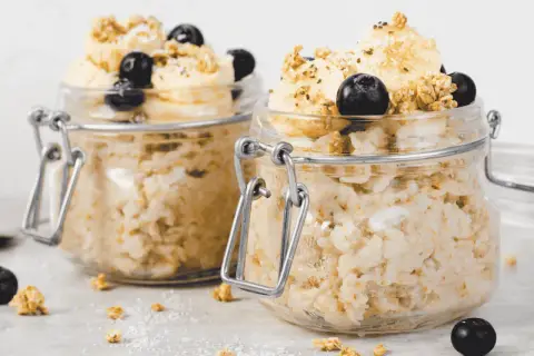 Zwei Gläser mit Overnight Oats, garniert mit Bananenscheiben, Blaubeeren und Granola. Perfekt für ein schnelles Frühstück.
