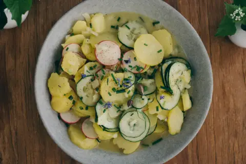 Bayrischer Kartoffelsalat mit Speck und Zwiebeln, garniert mit Radieschen, Gurken und Schnittlauch. Ein deftiger Genuss!