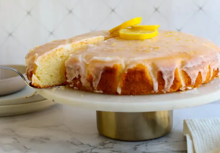 Ein saftiger Zitronenkuchen mit Zuckerguss und Zitronenscheiben, der beste Zitronenkuchen der Welt, wird auf einer Tortenplatte präsentiert.