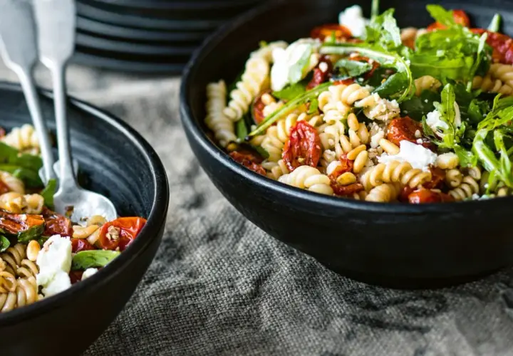 Zwei Schalen mit Nudelsalat mit getrockneten Tomaten, Pinienkernen und Rucola. Ein Löffel liegt in der linken Schale.
