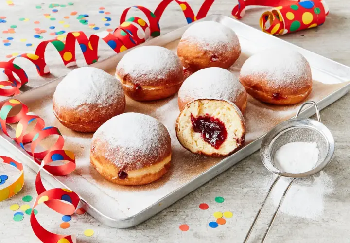 Berliner Rezept Original: Sieben luftige Krapfen mit Marmeladenfüllung, bestäubt mit Puderzucker, auf einem Blech, umgeben von bunten Party-Girlanden und Konfetti.