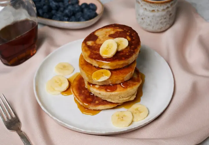Ein Stapel goldbrauner Bananen-Pancakes, beträufelt mit Sirup und garniert mit Bananenscheiben. Daneben Blaubeeren, Ahornsirup und Kaffee.