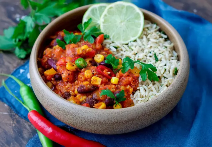 Herzhaftes Chili sin Carne mit Reis, garniert mit frischer Petersilie und Limettenspalten. Rote und grüne Chilis liegen daneben.