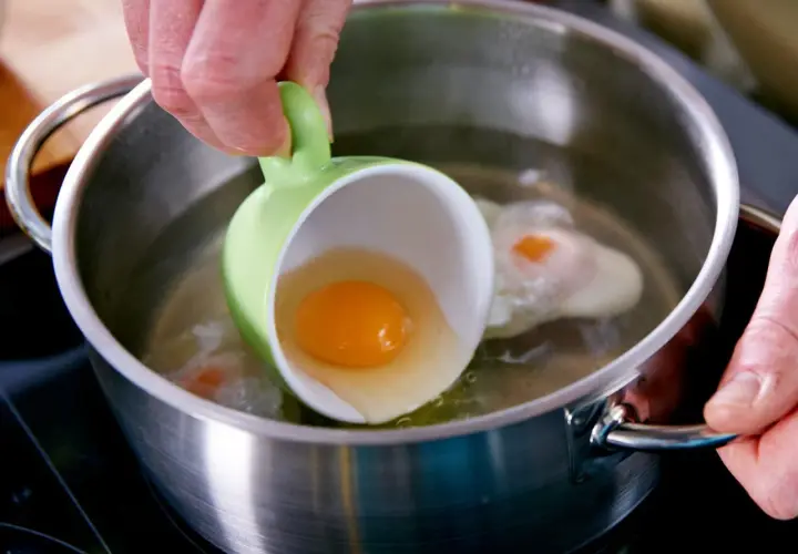 Ein rohes Ei wird aus einer grünen Tasse in kochendes Wasser gegossen. Ein Schritt für ein perfektes verlorene Eier Rezept.