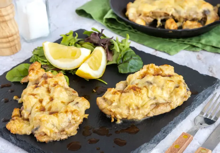 Zwei Portionen überbackenes Steak au four mit geschmolzenem Käse, serviert mit Salat und Zitronenspalten auf einem schwarzen Schiefer.