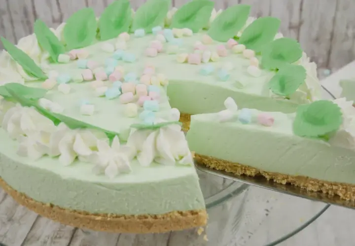 Eine helle, cremige Philadelphia Torte mit Waldmeistergeschmack, dekoriert mit Marshmallows und grünen Blättern. Ein Stück wird gerade serviert.