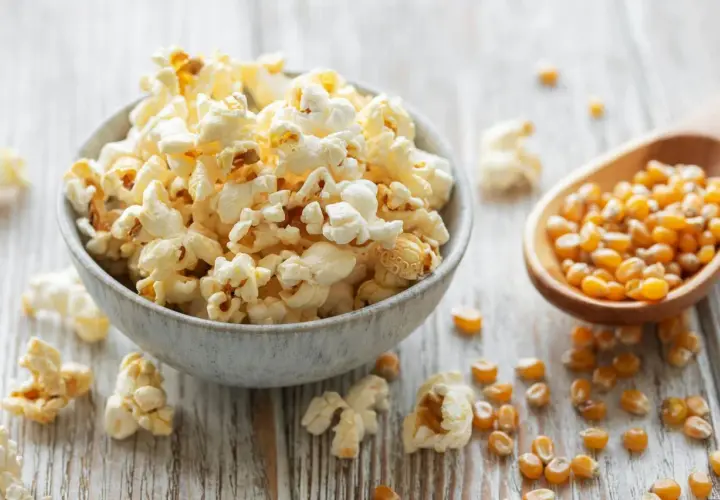 Schüssel mit frisch gemachtem Popcorn, daneben Maiskörner in einem Holzlöffel. So einfach ist Popcorn selber machen!