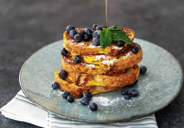 Ein Stapel goldbrauner French Toast, garniert mit Blaubeeren und Puderzucker. Ein dünner Strahl Sirup fließt darüber.