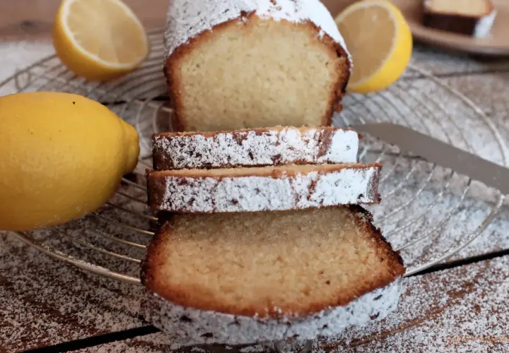 Zitronenkuchen wie bei Oma, mit Puderzucker bestäubt, neben frischen Zitronen auf einem Gitterrost.