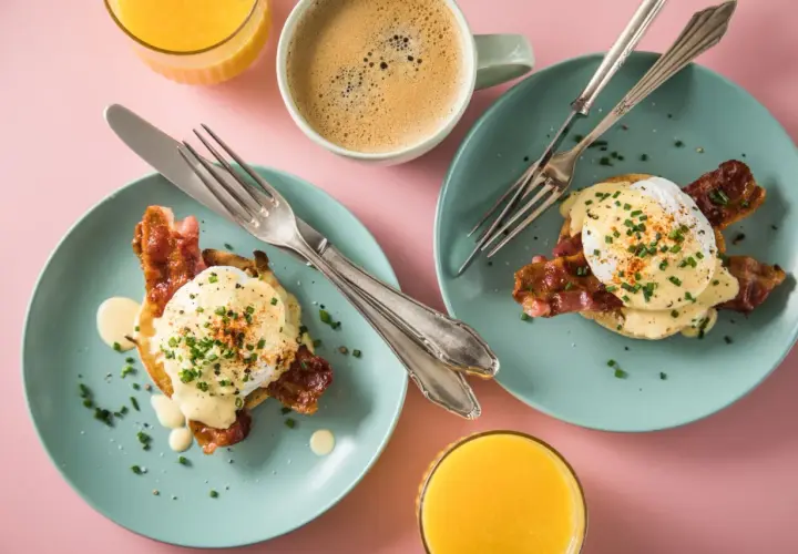 Zwei Teller mit Eggs Benedict, dazu Kaffee und Orangensaft auf rosa Untergrund.