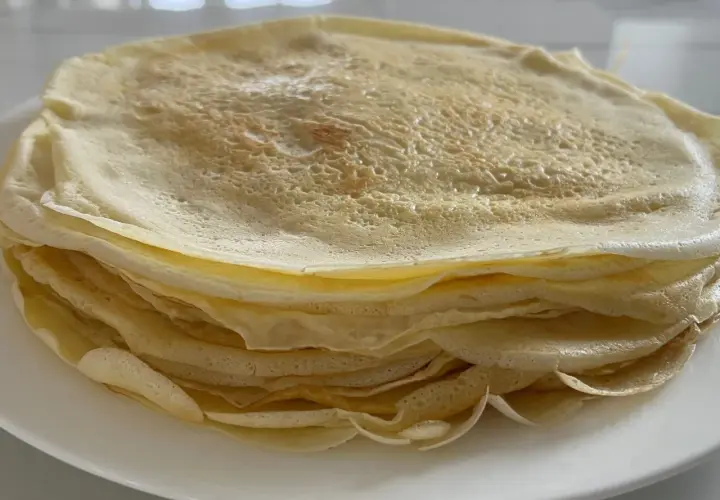 Ein Stapel goldbrauner Crêpes, frisch zubereitet aus dem Teig, liegt auf einem weißen Teller.