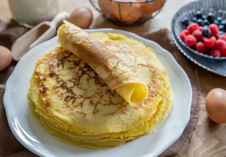 Ein Stapel goldbrauner Pfannkuchen, einer davon aufgerollt, wartet auf ein leckeres Pfannkuchen Rezept. Beeren und Eier im Hintergrund.