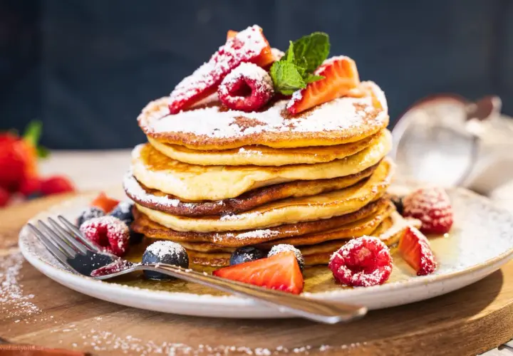Ein Stapel fluffiger amerikanischer Pancakes, bestäubt mit Puderzucker und garniert mit frischen Beeren und Minze. Ein köstliches american pancakes rezept für den perfekten Brunch.