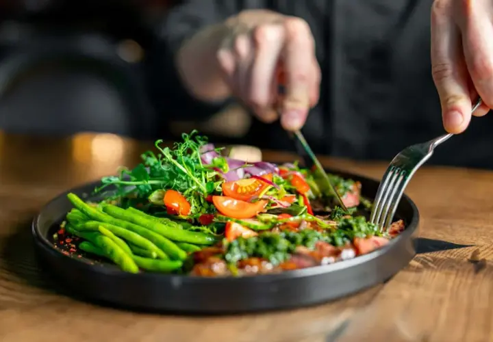 Ein Teller mit frischem Gemüse, wie grüne Bohnen und Tomaten, inspiriert von clean eating rezepten. Eine Person schneidet das Essen mit Messer und Gabel.