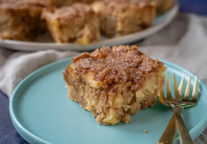Ein Stück saftiger Apfel Zimt Kuchen mit knuspriger Kruste auf einem türkisfarbenen Teller, daneben zwei goldene Gabeln.
