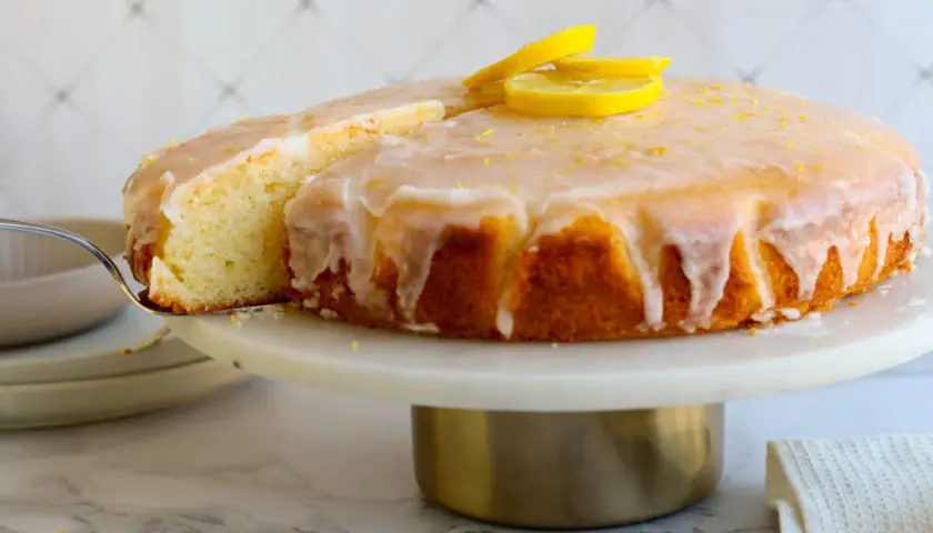 Ein saftiger Zitronenkuchen mit Zuckerguss und Zitronenscheiben, der beste Zitronenkuchen der Welt, wird auf einer Tortenplatte präsentiert.