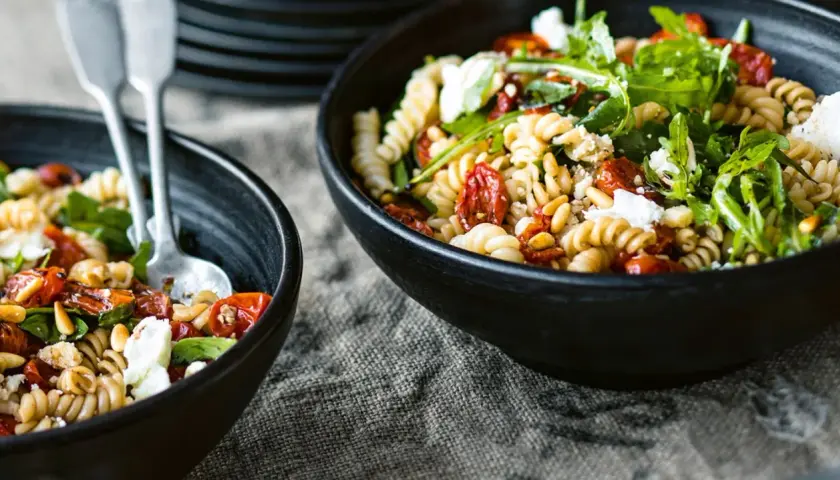 Zwei Schalen mit Nudelsalat mit getrockneten Tomaten, Pinienkernen und Rucola. Ein Löffel liegt in der linken Schale.