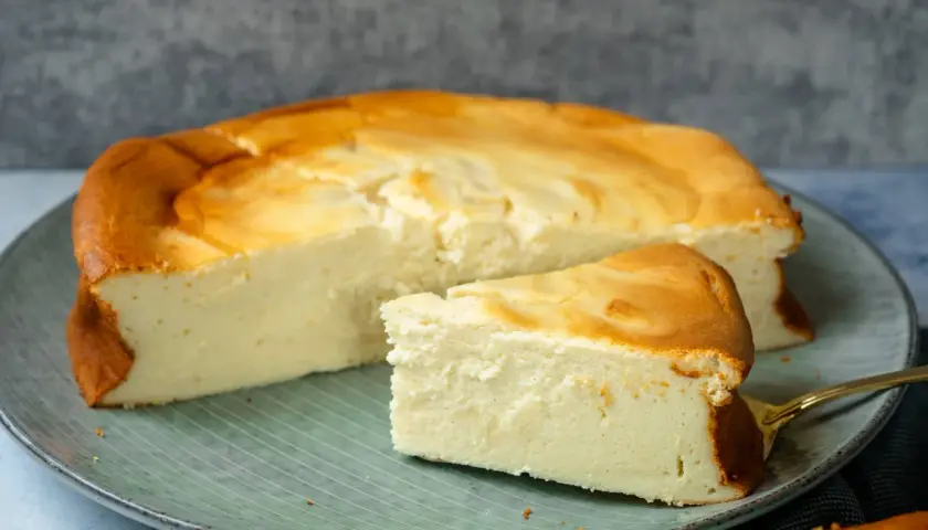 Ein saftiger Quarkkuchen ohne Boden, goldbraun gebacken, liegt auf einem Teller. Ein Stück ist bereits angeschnitten.
