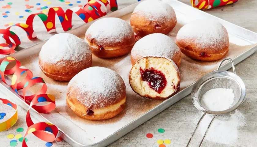 Berliner Rezept Original: Sieben luftige Krapfen mit Marmeladenfüllung, bestäubt mit Puderzucker, auf einem Blech, umgeben von bunten Party-Girlanden und Konfetti.