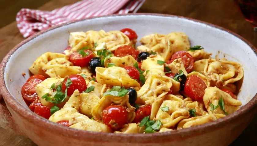 Herzhafter Tortellini Salat mit Kirschtomaten, schwarzen Oliven und frischem Basilikum in einer rustikalen Schüssel.