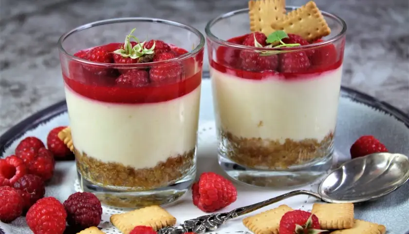 Zwei Gläser mit Himbeer-Dessert: eine Schicht Keksbrösel, cremige Masse und frische Himbeeren mit Himbeersauce.
