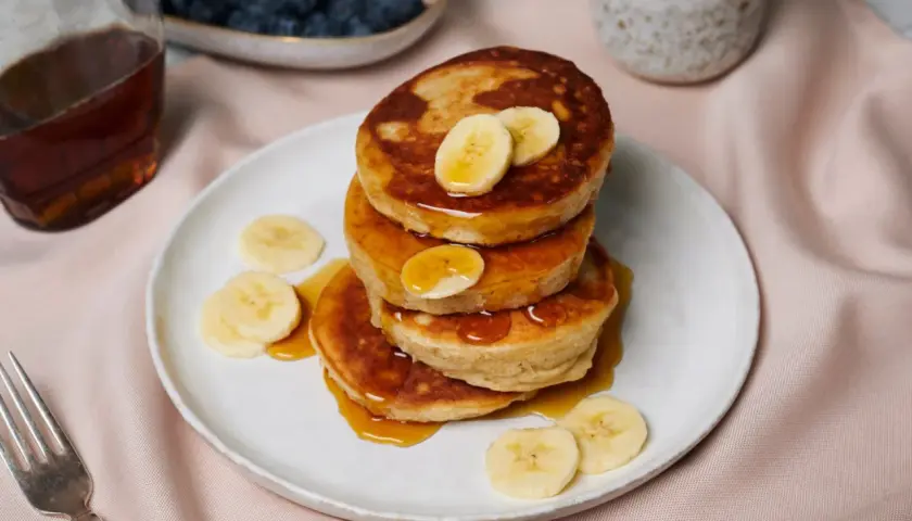 Ein Stapel goldbrauner Bananen-Pancakes, beträufelt mit Sirup und garniert mit Bananenscheiben. Daneben Blaubeeren, Ahornsirup und Kaffee.