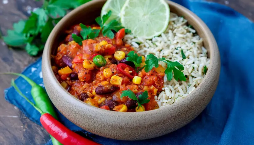 Herzhaftes Chili sin Carne mit Reis, garniert mit frischer Petersilie und Limettenspalten. Rote und grüne Chilis liegen daneben.