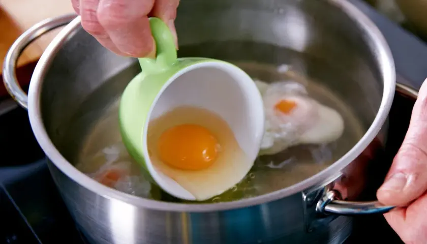 Ein rohes Ei wird aus einer grünen Tasse in kochendes Wasser gegossen. Ein Schritt für ein perfektes verlorene Eier Rezept.