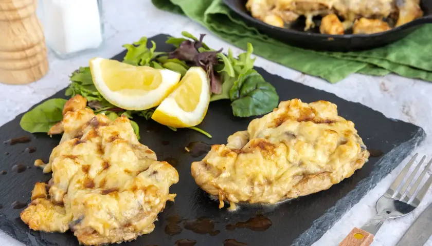 Zwei Portionen überbackenes Steak au four mit geschmolzenem Käse, serviert mit Salat und Zitronenspalten auf einem schwarzen Schiefer.