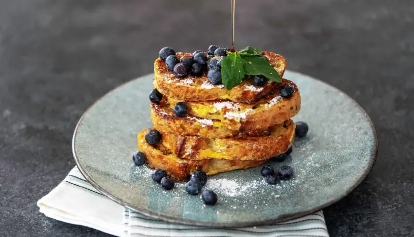 Ein Stapel goldbrauner French Toast, garniert mit Blaubeeren und Puderzucker. Ein dünner Strahl Sirup fließt darüber.