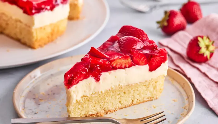 Ein saftiges Stück Erdbeer Schmand Kuchen mit frischen Erdbeeren und Tortenguss auf einem Teller, daneben frische Erdbeeren und eine Gabel.