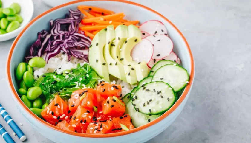 Bunte Poke Bowl mit Lachs, Avocado, Edamame, Gurke, Radieschen, Rotkohl und Seetang auf Reis.