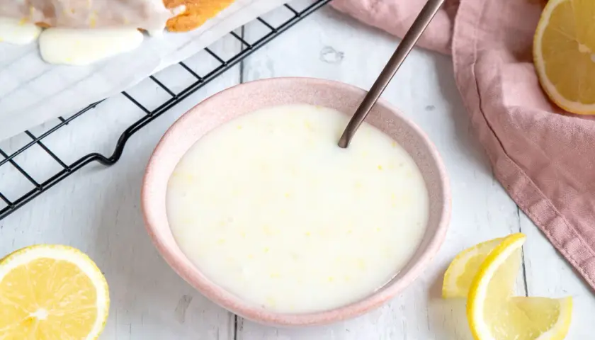 Zitronenglasur selber machen: cremige Glasur in einer Schale, daneben Zitronenspalten und ein Stück Kuchen auf einem Gitter.